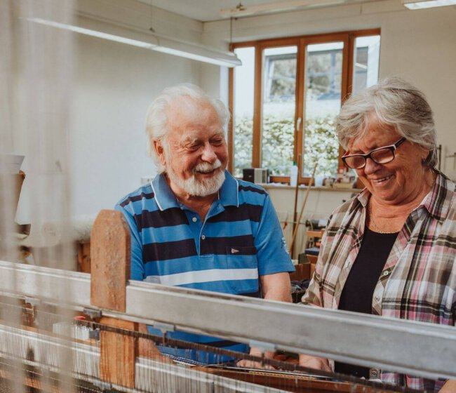Zwei ältere Personen arbeiten gemeinsam an einem Webstuhl.